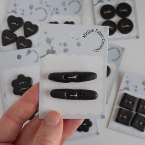 Peut inclure: Divers boutons en céramique noire de formes variées : cœurs, cercles, carrés et fleurs. Les boutons sont présentés sur des cartes blanches avec le texte "Mirjam Faber Ceramics".