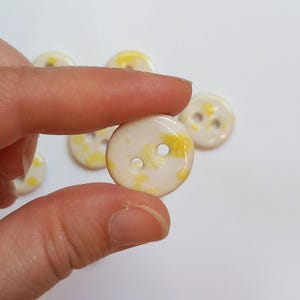 Può includere: Un set di sei bottoni in ceramica bianca con macchie gialle. Ogni bottone ha due fori.