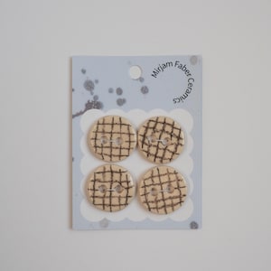 Peut inclure: Quatre boutons ronds en céramique, beige avec un motif quadrillé marron foncé. Les boutons sont fixés sur une carte bleu clair avec le texte "mirjam faber ceramics". Les boutons ont deux trous pour la couture.