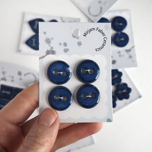 Può includere: Quattro bottoni rotondi in ceramica blu scuro con finitura lucida e due fori al centro, fissati su un cartoncino bianco con l'etichetta Mirjam Faber Ceramics. I bottoni presentano una superficie liscia, riflettente e un colore blu uniforme.