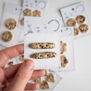 Puede incluir: Una mano sostiene una tarjeta con dos botones de cerámica moteada. Los botones son de color beige con manchas marrones y tienen dos agujeros. En el fondo hay otras tarjetas con botones de cerámica de varias formas y tamaños. La tarjeta dice "Mirjam Faber Ceramics."