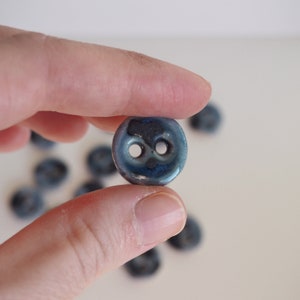 May include: A small, round, two-hole button with a glossy, variegated blue and dark grey glaze. The surface features a smooth, reflective finish with subtle color variations blending from deep navy to lighter shades of blue across the circular form.