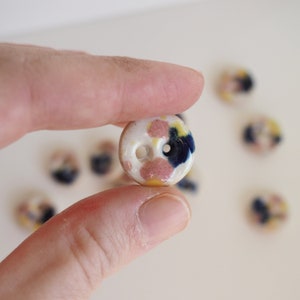 Könnte beinhalten: Nahaufnahme eines Keramikknopfes mit weißer Basis und rosa, gelben und blauen Flecken. Der Knopf hat zwei Löcher.
