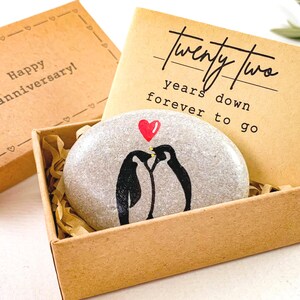 May include: A gray stone with two black penguins kissing under a red heart, presented in a brown box with the text "Twenty Two years down forever to go." The box is accompanied by a separate box with the text "Happy anniversary!"