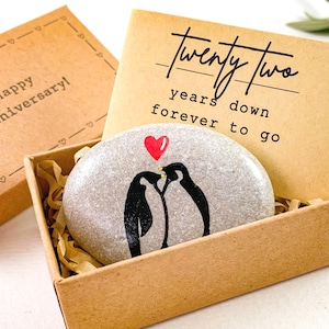 May include: A gift box with a painted stone featuring two penguins under a red heart. The stone is nestled in a cardboard box with the words "Twenty Two years down forever to go" on a card. The box also includes a separate lid with the words "Happy Anniversary!"