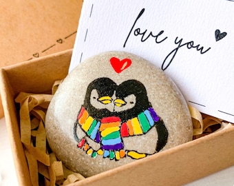 Puede incluir: Una piedra pintada con dos pingüinos que llevan una bufanda arcoíris, un corazón rojo sobre ellos y las palabras "love you" en una tarjeta en una pequeña caja marrón.