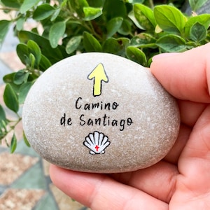 May include: A smooth, gray stone with the words "Camino de Santiago" in black script, an upward yellow arrow, and a shell symbol. The stone is held in a hand against a backdrop of green leaves.
