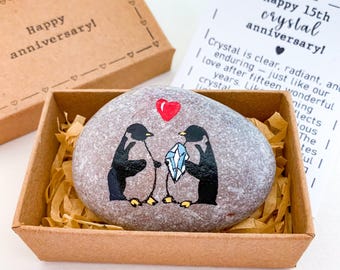 May include: A painted pebble featuring two penguins presenting a crystal, nestled in a cardboard box with paper filling. A red heart is painted above. The box lid says "Happy anniversary!" A card reads "Happy 15th crystal anniversary!"