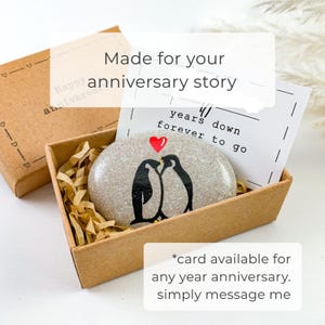May include: A gift box containing a painted stone with two black penguins kissing under a red heart. The stone is in a box with shredded paper and a card that says "years down forever to go". The box also has the text "Happy anniversary".