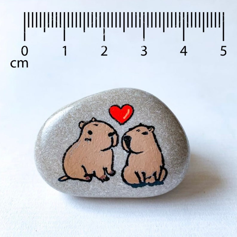 May include: A hand-painted, gray stone featuring two capybaras with a red heart above them. The capybaras are brown with black outlines. The stone is approximately 4.5 cm long.