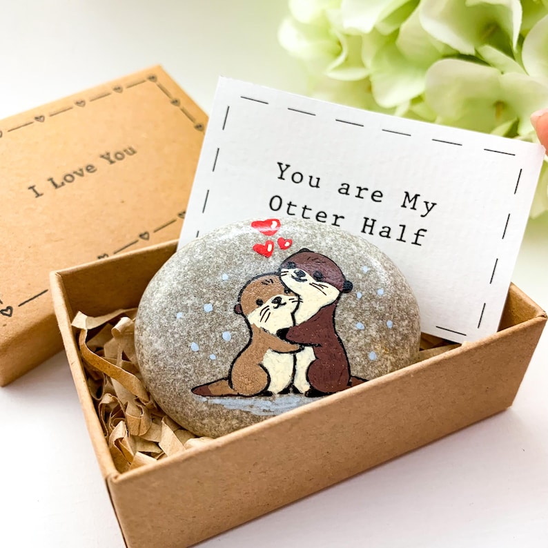 Pu&ograve; includere: Un sasso dipinto con due lontre che si abbracciano, con cuori rossi sopra, all'interno di una scatola marrone. Una carta con la scritta "You are My Otter Half" &egrave; dietro la pietra. La scatola &egrave; in una scatola pi&ugrave; grande con "I Love You".