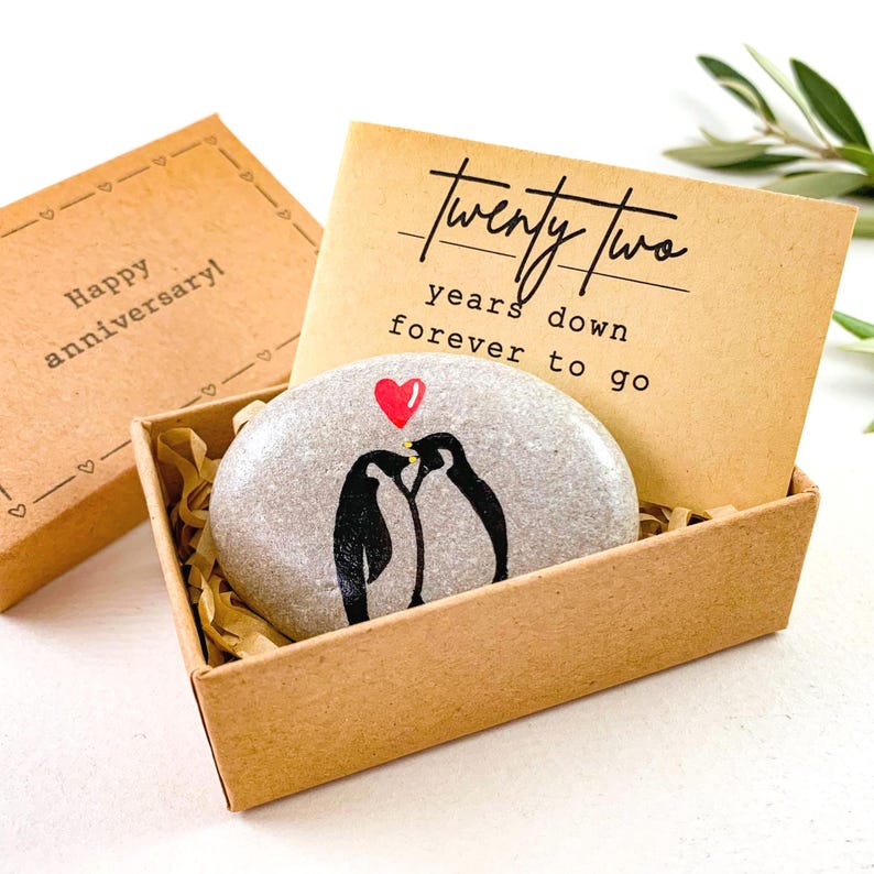 May include: A gift box with a painted stone featuring two penguins under a red heart. The stone is nestled in a cardboard box with the words "Twenty Two years down forever to go" on a card. The box also includes a separate lid with the words "Happy Anniversary!"