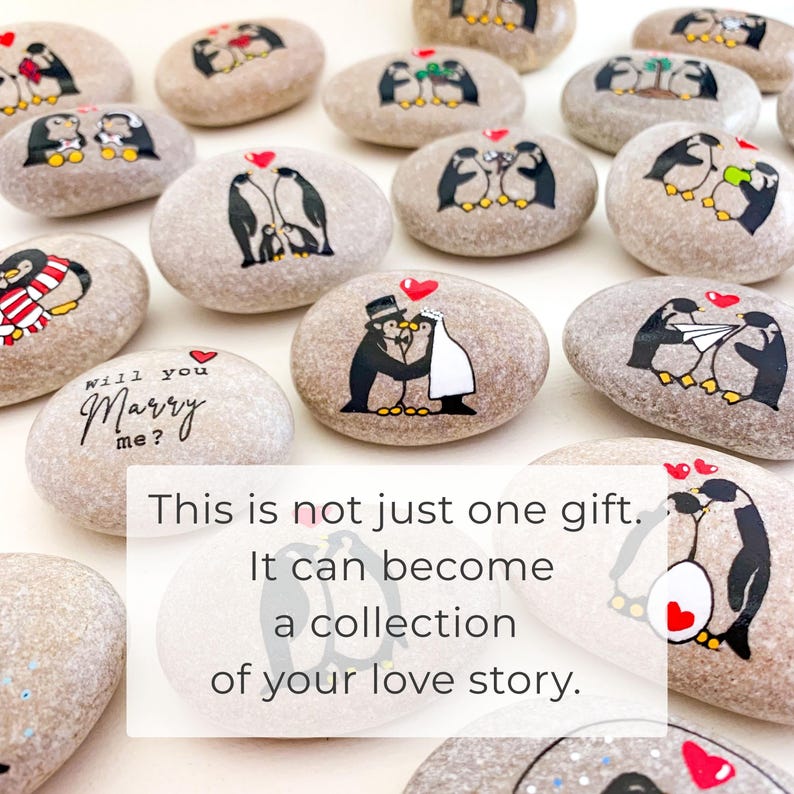 May include: A collection of painted stones featuring penguin illustrations. Some stones depict penguins in wedding attire, others with hearts, and some with the phrase "Will you Marry me?". The text on the image reads: "This is not just one gift. It can become a collection of your love story."