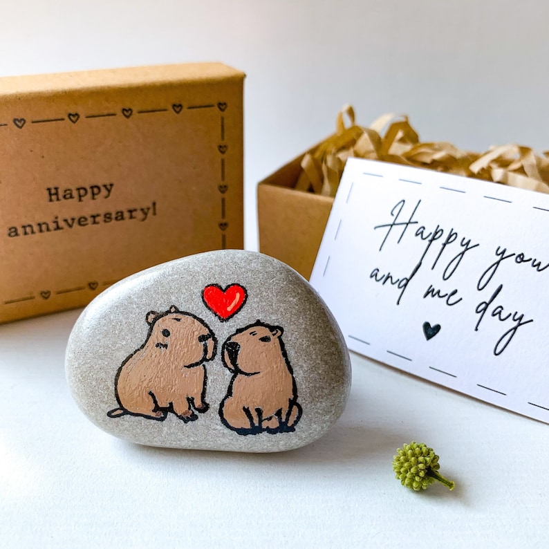 May include: A painted stone featuring two capybaras with a red heart above them. The stone is accompanied by a gift box with the text "Happy anniversary!" and a card that reads "Happy you and me day".