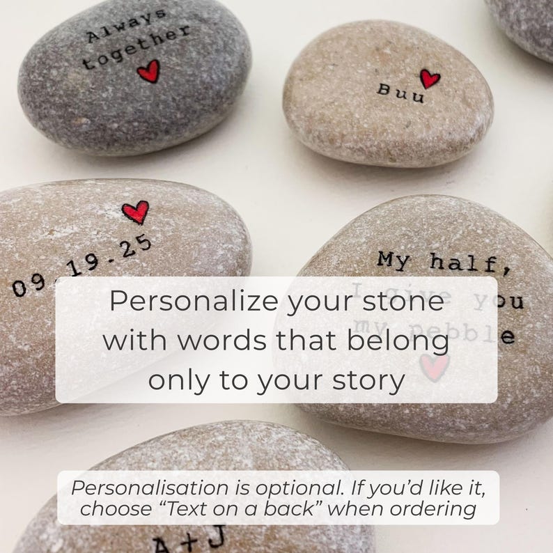 May include: A collection of smooth, gray and tan river stones, each inscribed with personalized text and a small red heart. The stones feature phrases like "Always together" and "My half," with the option for custom text.