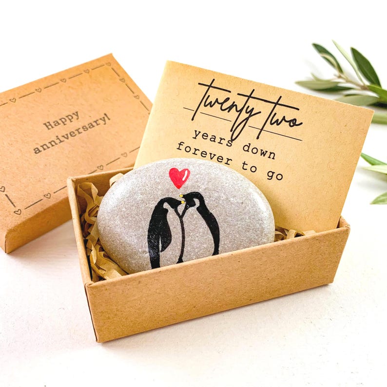 May include: A gray stone with two black penguins kissing under a red heart, presented in a brown box with the text "Twenty Two years down forever to go." The box is accompanied by a separate box with the text "Happy anniversary!"