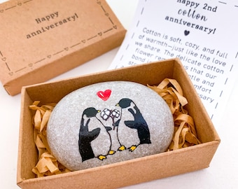 May include: A painted stone with two penguins holding flowers, inside a cardboard box with shredded paper. The stone is inscribed with a red heart. The box lid says "Happy anniversary!" and a card says "Happy 2nd cotton anniversary!"
