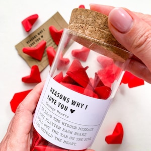May include: A glass jar filled with red paper hearts, each with a hidden message inside. The jar has a white label that reads "Reasons Why I Love You" with a small heart icon. The label also includes instructions on how to read the messages: "To read the hidden message, just flatten each heart, pull out the tab on the side, and unfold the heart."