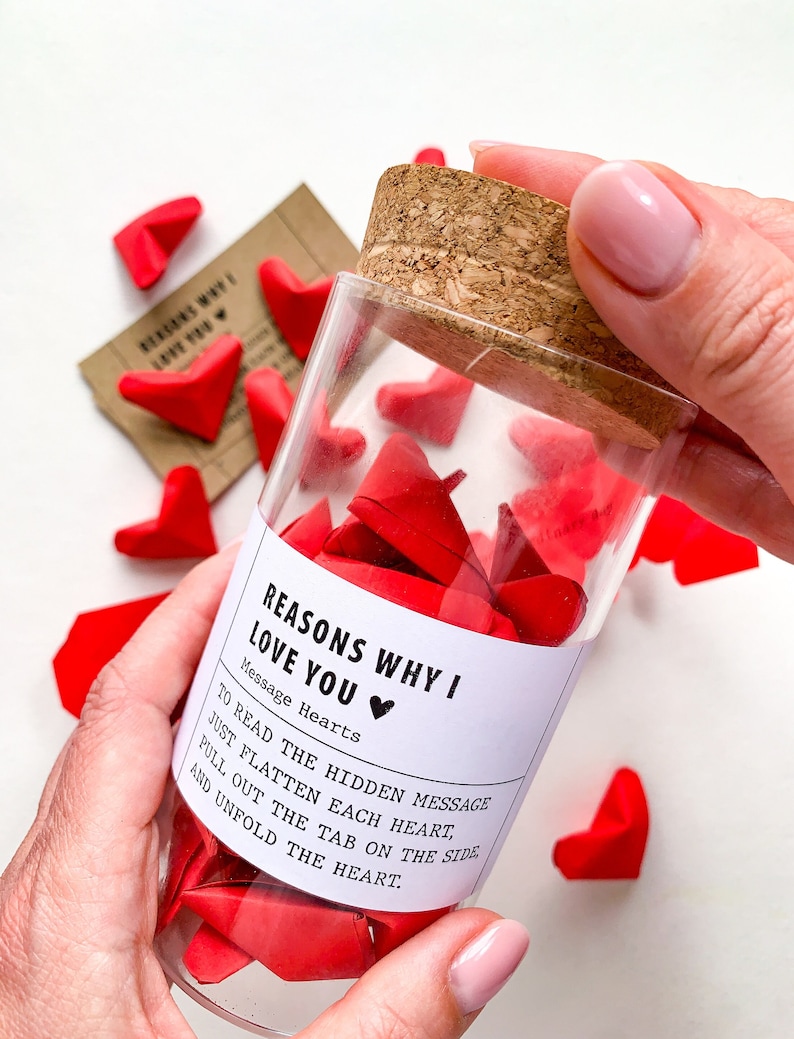 May include: A glass jar filled with red paper hearts, each with a hidden message inside. The jar has a white label that reads "Reasons Why I Love You" with a small heart icon. The label also includes instructions on how to read the messages: "To read the hidden message, just flatten each heart, pull out the tab on the side, and unfold the heart."