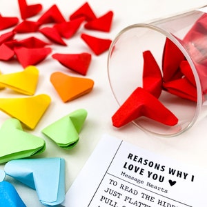 May include: A white card with the text "Reasons why I love you" and "Message Hearts" with a red heart emoji.  The card is next to a glass jar filled with colorful origami hearts.