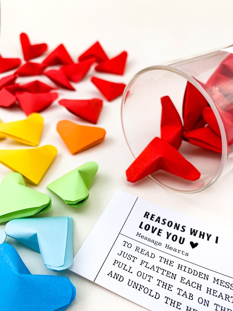 May include: A white card with the text "Reasons why I love you" and "Message Hearts" with a red heart emoji.  The card is next to a glass jar filled with colorful origami hearts.
