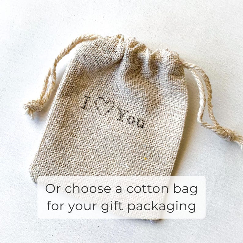 May include: A small, beige drawstring bag with the words "I ♡ You" printed on the front. The bag is made of a textured fabric and has a natural, rustic appearance. The text "Or choose a cotton bag for your gift packaging" is also visible.