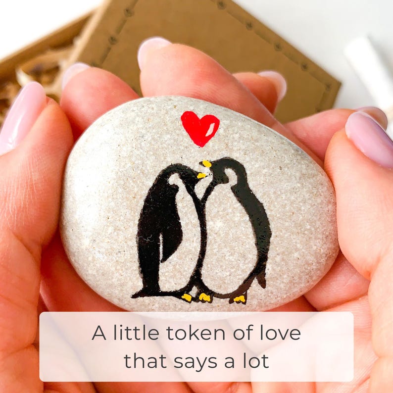 May include: A painted stone with two black penguins kissing under a red heart. The stone is held in a person's hands. Text on the bottom reads: "A little token of love that says a lot."