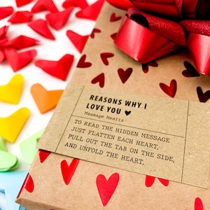 May include: A brown gift box wrapped in paper with red hearts printed on it. A red satin ribbon is tied around the box in a bow. A label on the box reads "Reasons Why I Love You" and "Message Hearts". The label also includes instructions on how to read the hidden message inside the hearts.