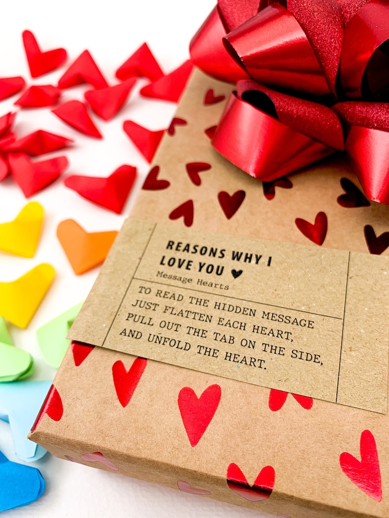 May include: A brown gift box wrapped in paper with red hearts printed on it. A red satin ribbon is tied around the box in a bow. A label on the box reads "Reasons Why I Love You" and "Message Hearts". The label also includes instructions on how to read the hidden message inside the hearts.