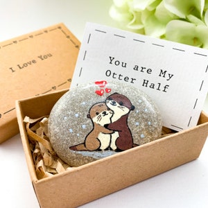Op de afbeelding: Een kleine, grijze steen beschilderd met twee otters die elkaar omhelzen. De steen zit in een klein, bruin doosje met een wit kaartje waarop staat "You are My Otter Half".