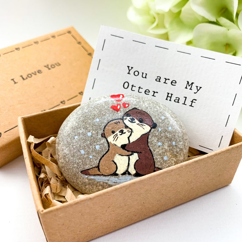 Op de afbeelding: Een kleine, grijze steen beschilderd met twee otters die elkaar omhelzen. De steen zit in een klein, bruin doosje met een wit kaartje waarop staat "You are My Otter Half".