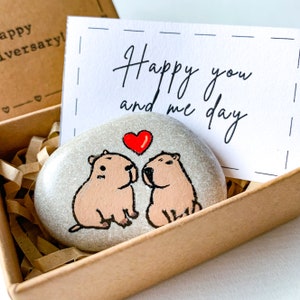 May include: A painted stone featuring two capybaras with a red heart above them, inside a wooden box with shredded paper. A card with the text "Happy you and me day" is behind the stone.
