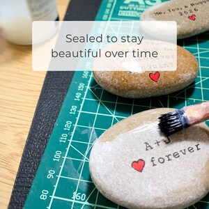 May include: Close-up of painted rocks with text and red heart accents. One rock reads "A+J forever" and another reads "Me, You, & Hugs 2026". A paintbrush is applying a sealant. The text "Sealed to stay beautiful over time" is also present.