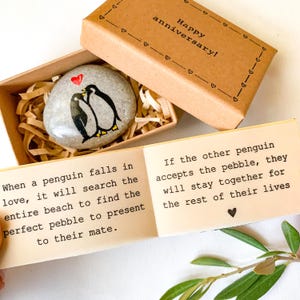 May include: A painted stone with two penguins and a red heart, presented in a small box with the words "Happy anniversary!". The box is open, revealing the stone nestled in shredded paper. A card with text about penguins is also visible.