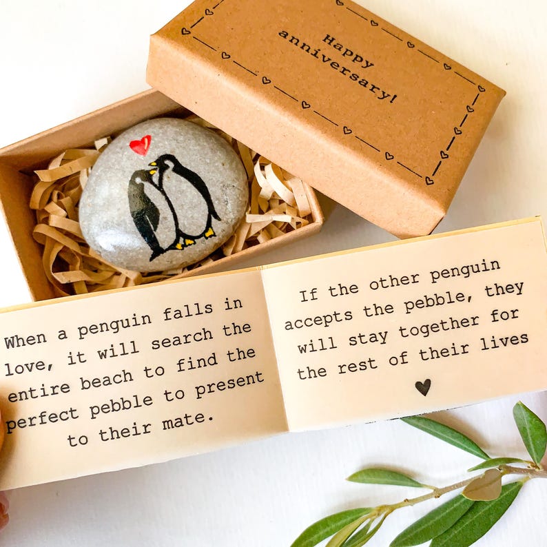 May include: A painted stone with two penguins and a red heart, presented in a small box with the words "Happy anniversary!". The box is open, revealing the stone nestled in shredded paper. A card with text about penguins is also visible.