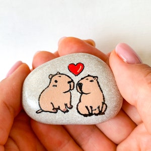 May include: A hand-painted, light gray stone featuring two capybaras facing each other, with a red heart above them. The capybaras are painted in shades of brown and black, creating a charming, whimsical design.