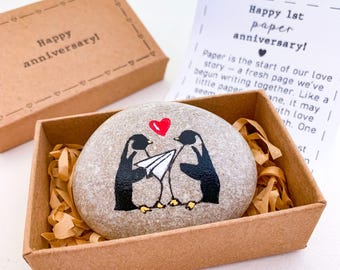 May include: A painted stone featuring two penguins with a paper airplane and a red heart, with the text "Happy 1st paper anniversary!" in a brown box with paper. Another box with "Happy anniversary!" is also visible.