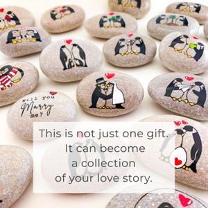 May include: A collection of painted stones featuring penguin illustrations. Some stones depict penguins in wedding attire, others with hearts, and some with the phrase "Will you Marry me?". The text on the image reads: "This is not just one gift. It can become a collection of your love story."
