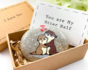 May include: A small, gray stone painted with two otters hugging. The otters are brown and white. The stone is in a small, brown box with a white card that says "You are my Otter Half".