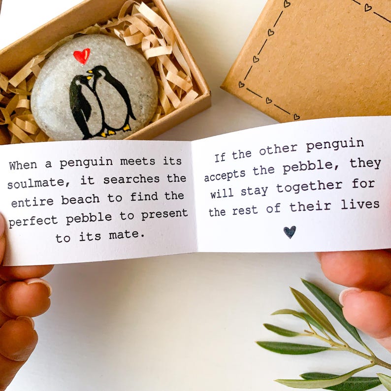 May include: A painted pebble in a gift box, featuring two penguins kissing under a red heart. The box contains a small card with text about penguins and soulmates. A second box with heart designs is also visible.