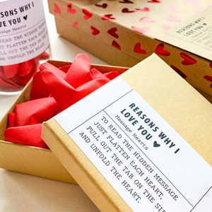 May include: A brown box with a white label that reads "Reasons Why I Love You" with a red heart. The label also includes instructions to read the hidden message by flattening each heart, pulling out the tab on the side, and unfolding the heart.