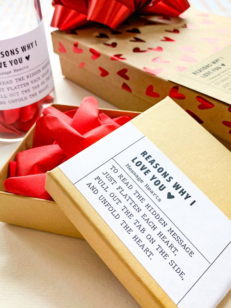 May include: A brown box with a white label that reads "Reasons Why I Love You" with a red heart. The label also includes instructions to read the hidden message by flattening each heart, pulling out the tab on the side, and unfolding the heart.