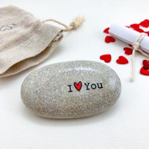 Może przedstawiać: Gładki, owalny, beżowy kamień rzeczny z czarnym napisem I, czerwonym symbolem serca i napisem YOU. Kamieniowi towarzyszy mały beżowy lniany woreczek ze sznurkiem oraz kilka rozsypanych małych czerwonych konfetti w kształcie serc.
