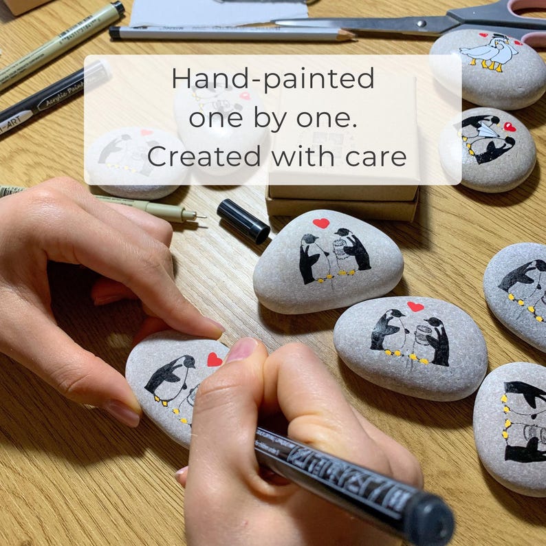 May include: Hand-painted, decorated stones with penguin designs and red hearts. The text on the image reads: "Hand-painted one by one. Created with care."