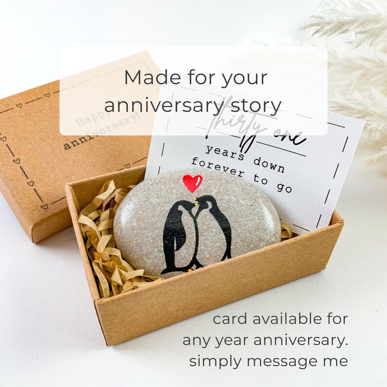 May include: A smooth gray stone painted with two black and white penguins and a small red heart, nestled in a brown cardboard gift box with shredded paper filler. A white card inside reads: thirty one years down forever to go.