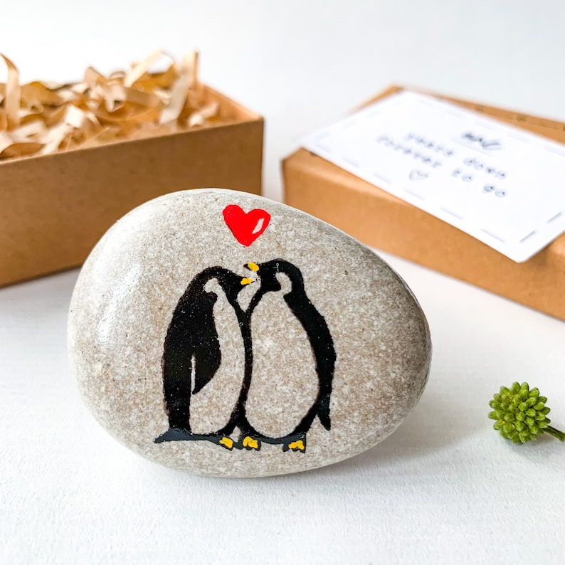 May include: A smooth, light gray river stone featuring a hand-painted illustration of two black and white penguins facing each other. Above the penguins is a small, solid red heart. The stone is oval-shaped with a natural, speckled texture.