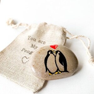May include: A painted stone featuring two black penguins with yellow beaks and feet, kissing under a red heart. A small beige drawstring bag with the text "You are My Peng" is in the background.