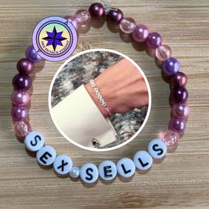 May include: A beaded bracelet with purple, pink, and clear beads. The bracelet spells out "SEX SELLS" with white letter beads. A circular inset shows a wrist wearing a similar bracelet.