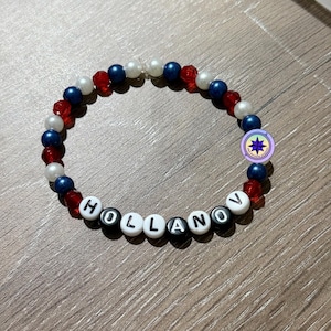 Op de afbeelding: Een kralenarmband met de naam "HOLLANOV" gespeld met witte letterkralen. De armband heeft rode, witte en blauwe kralen en een kleine ronde bedel. De armband ligt op een houten oppervlak.