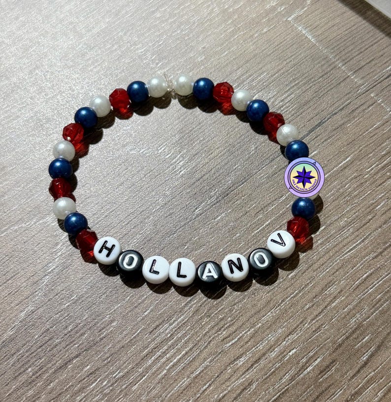 Op de afbeelding: Een kralenarmband met de naam "HOLLANOV" gespeld met witte letterkralen. De armband heeft rode, witte en blauwe kralen en een kleine ronde bedel. De armband ligt op een houten oppervlak.